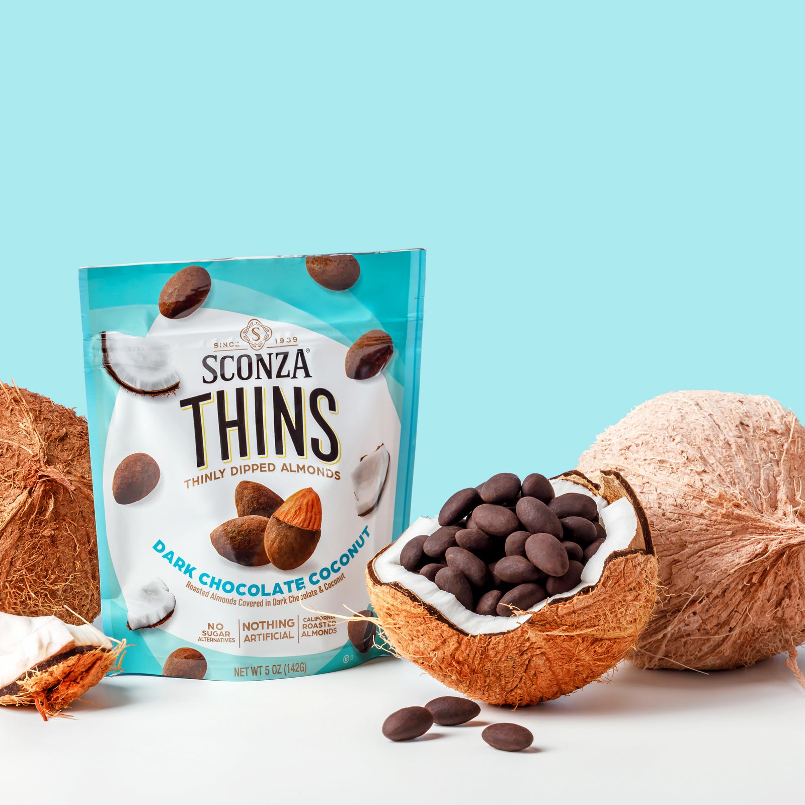 Sconza Thins dark chocolate coconut almonds with packaging and coconuts on a light blue background
