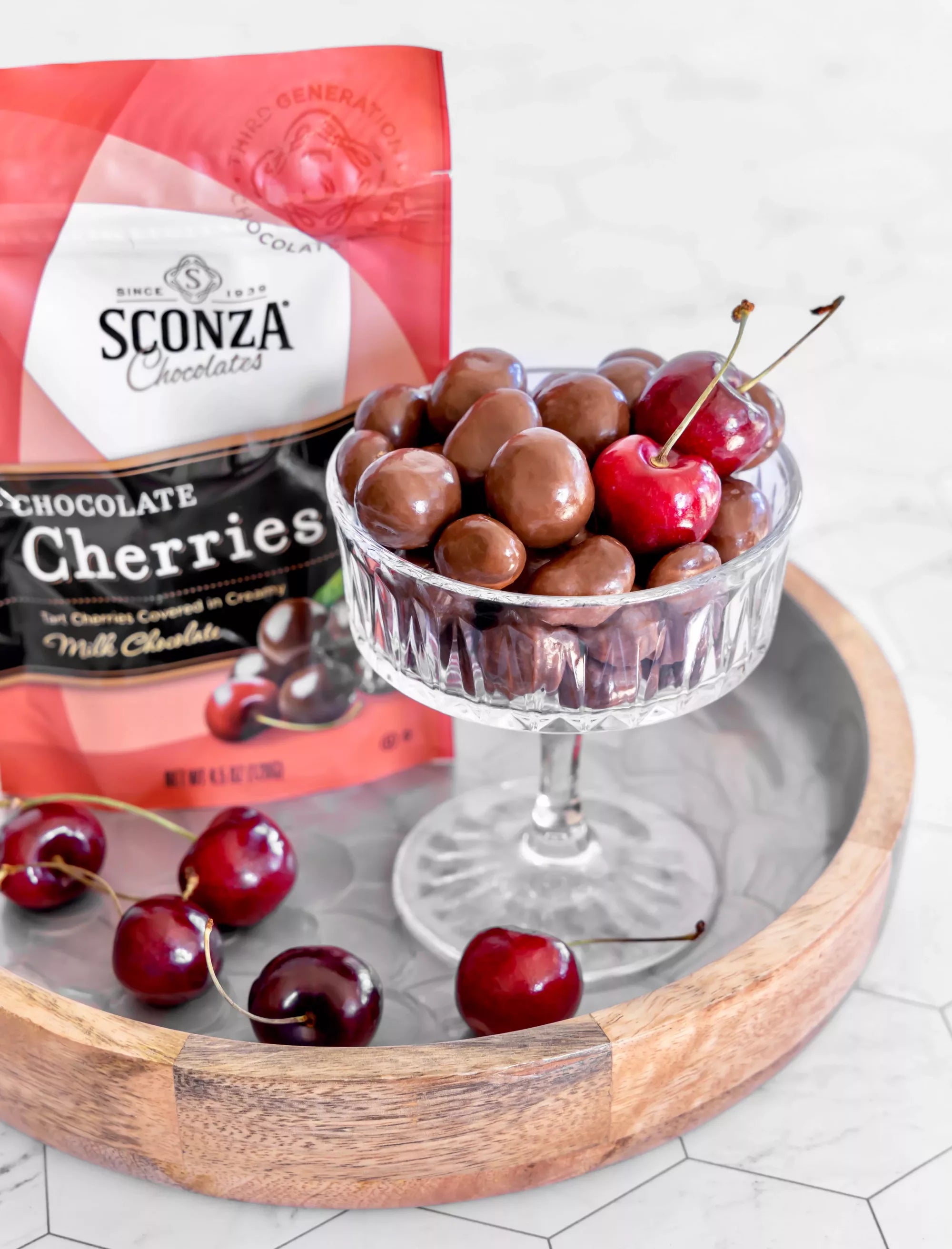 Sconza Chocolate covered cherries on a table, sitting in a glass dish surrounded by real cherries. 